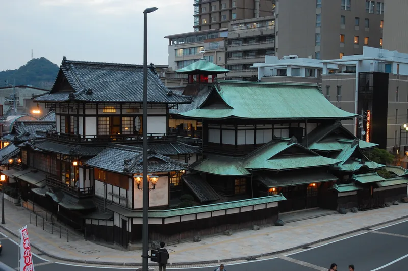Dōgo Onsen Honkan at dusk, with a tiled roof and illuminated windows.