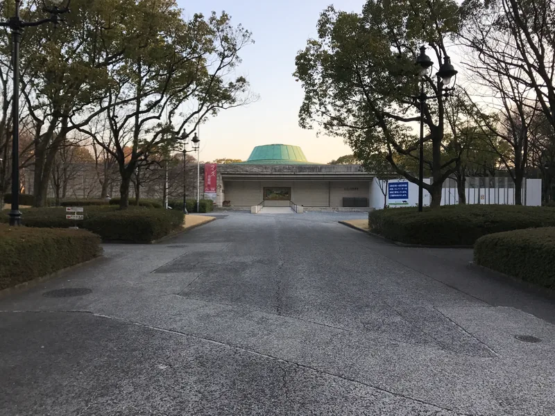 A tree-lined approach to Hiroshima Museum of Art, with a low facade and a green roof.